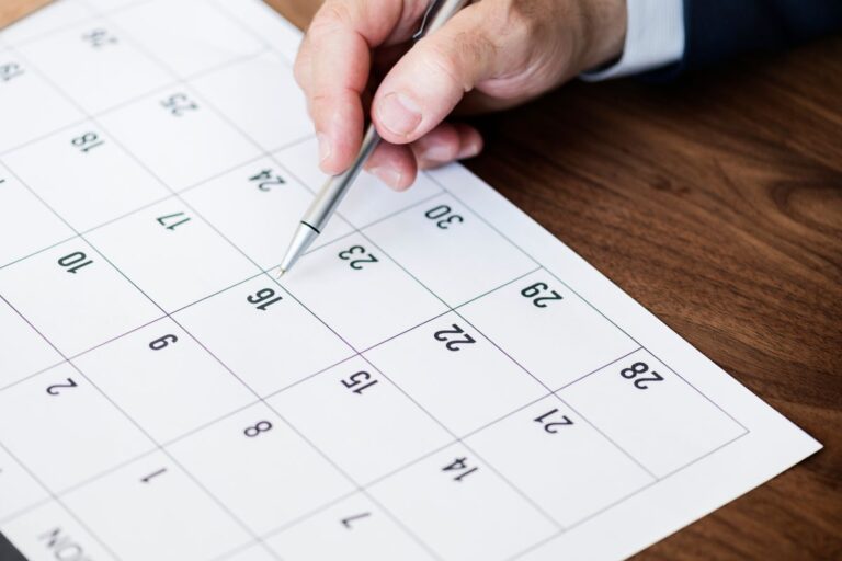 Plano geral de cima mostrando uma mão com uma caneta esferográfica de metal sobre um calendário de papel branco.