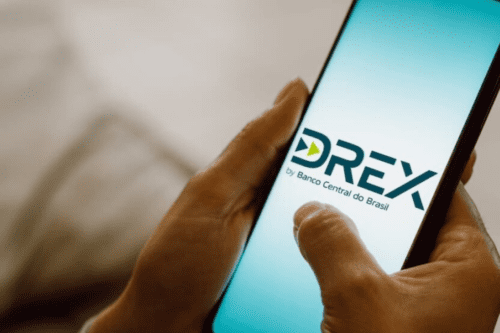 lose-up de mãos segurando um smartphone que exibe na tela o logotipo oficial do Drex, a moeda digital brasileira, com a inscrição 'by Banco Central do Brasil' logo abaixo
