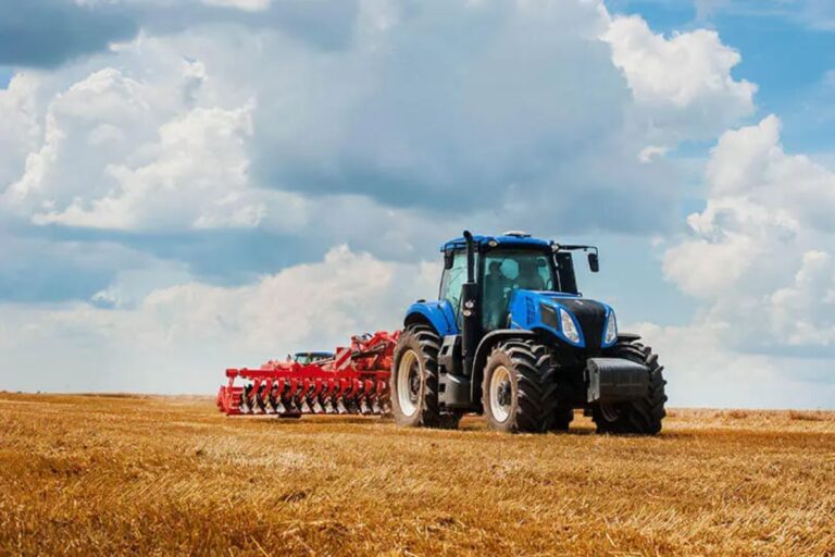 Trator azul de grande porte, puxando um implemento agrícola vermelho, trabalhando em um campo seco e dourado.