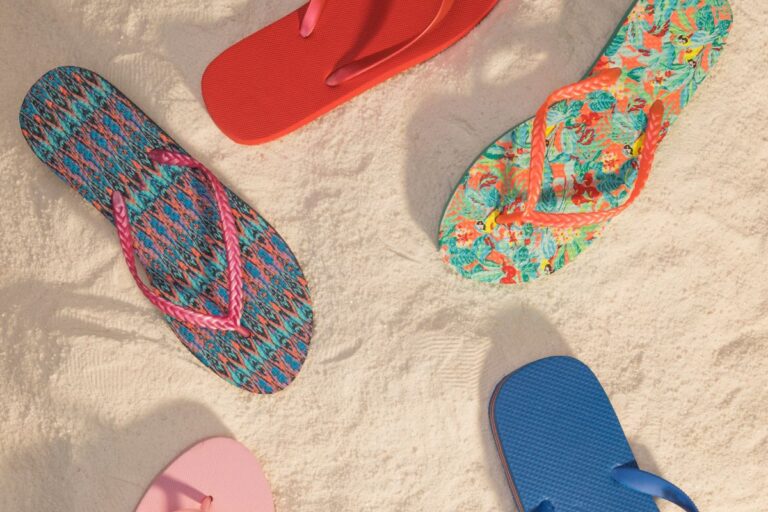 Vista aérea de cinco pares de chinelos dispostos sobre uma areia clara e fofa (praia ou dunas).