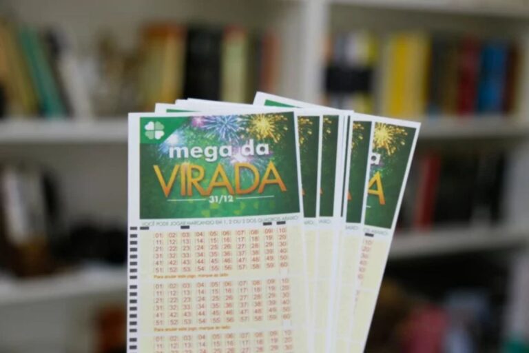 Mão segurando vários volantes de apostas da Mega da Virada contra um fundo desfocado de uma estante de livros. O bilhete é verde com detalhes em amarelo, exibindo o logotipo oficial do sorteio especial de 31 de dezembro e as tabelas numéricas para marcação.