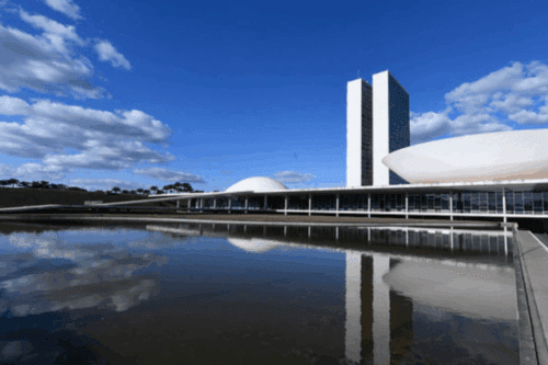 Vista panorâmica do Palácio do Congresso Nacional em Brasília sob um céu azul com nuvens. A imagem destaca as torres gêmeas e as duas cúpulas icônicas refletidas na clareza do espelho d'água em primeiro plano.