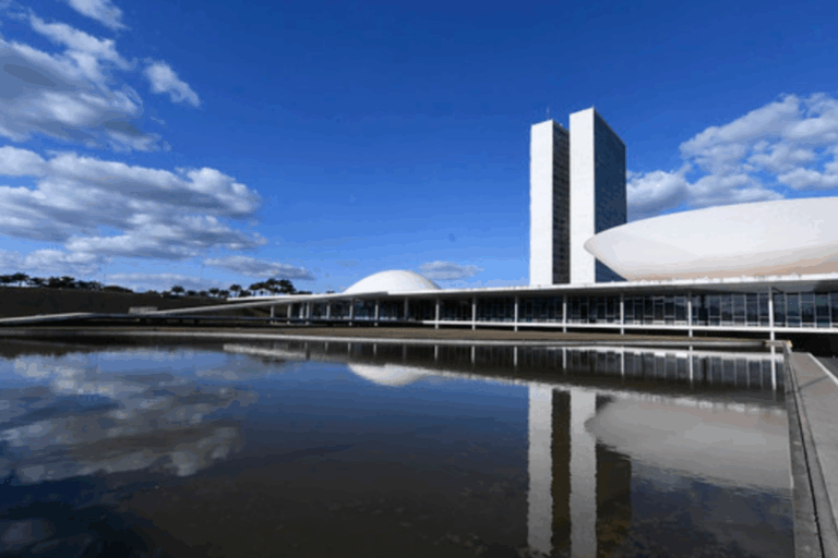 Vista panorâmica do Palácio do Congresso Nacional em Brasília sob um céu azul com nuvens. A imagem destaca as torres gêmeas e as duas cúpulas icônicas refletidas na clareza do espelho d'água em primeiro plano.