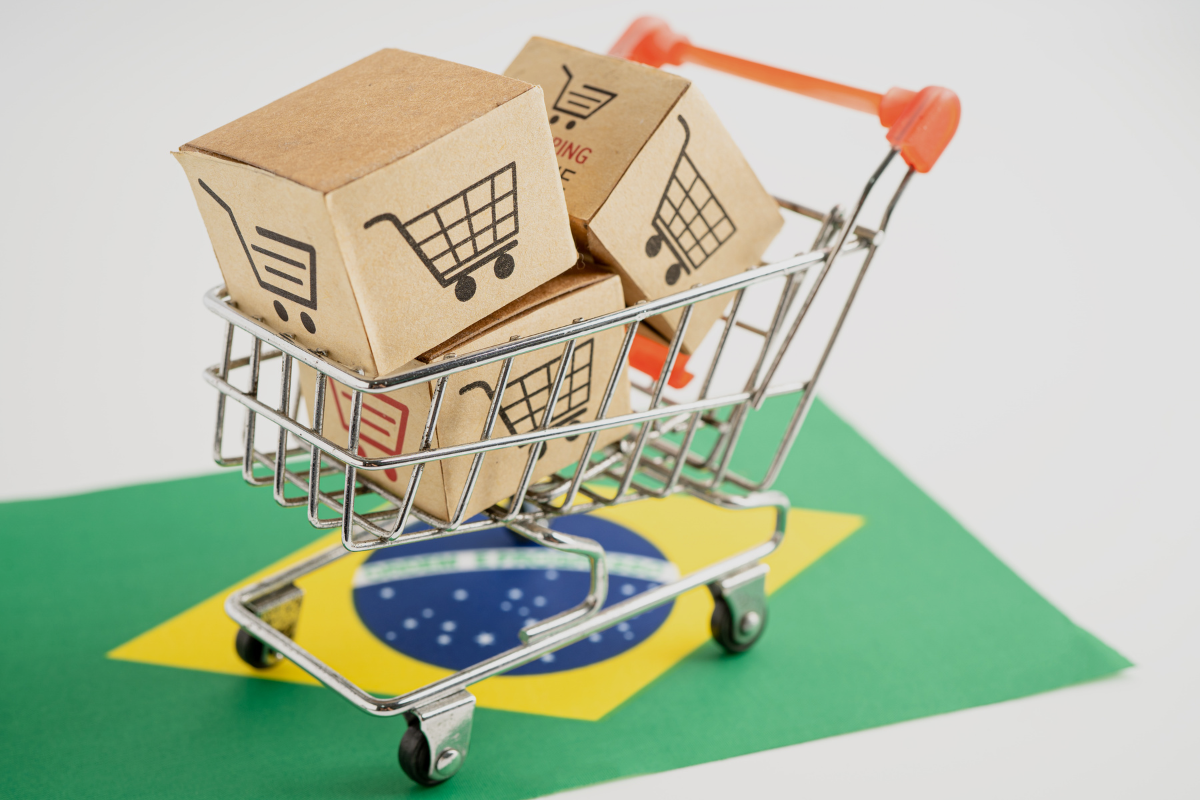 Carrinho de supermercado com caixas de papelão contendo símbolos de carrinho de compras, representando compras online no Brasil, com mapa do Brasil ao fundo.